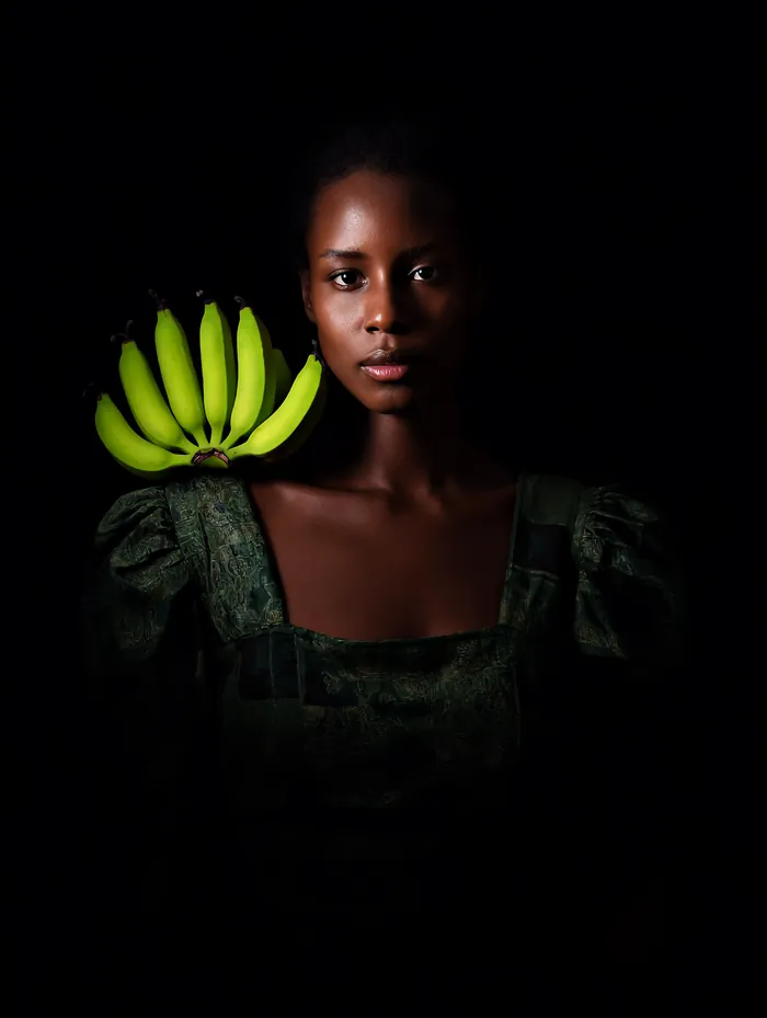 A Ugandan woman in a gomesi holding a hand of green matooke beside her face, lit against black